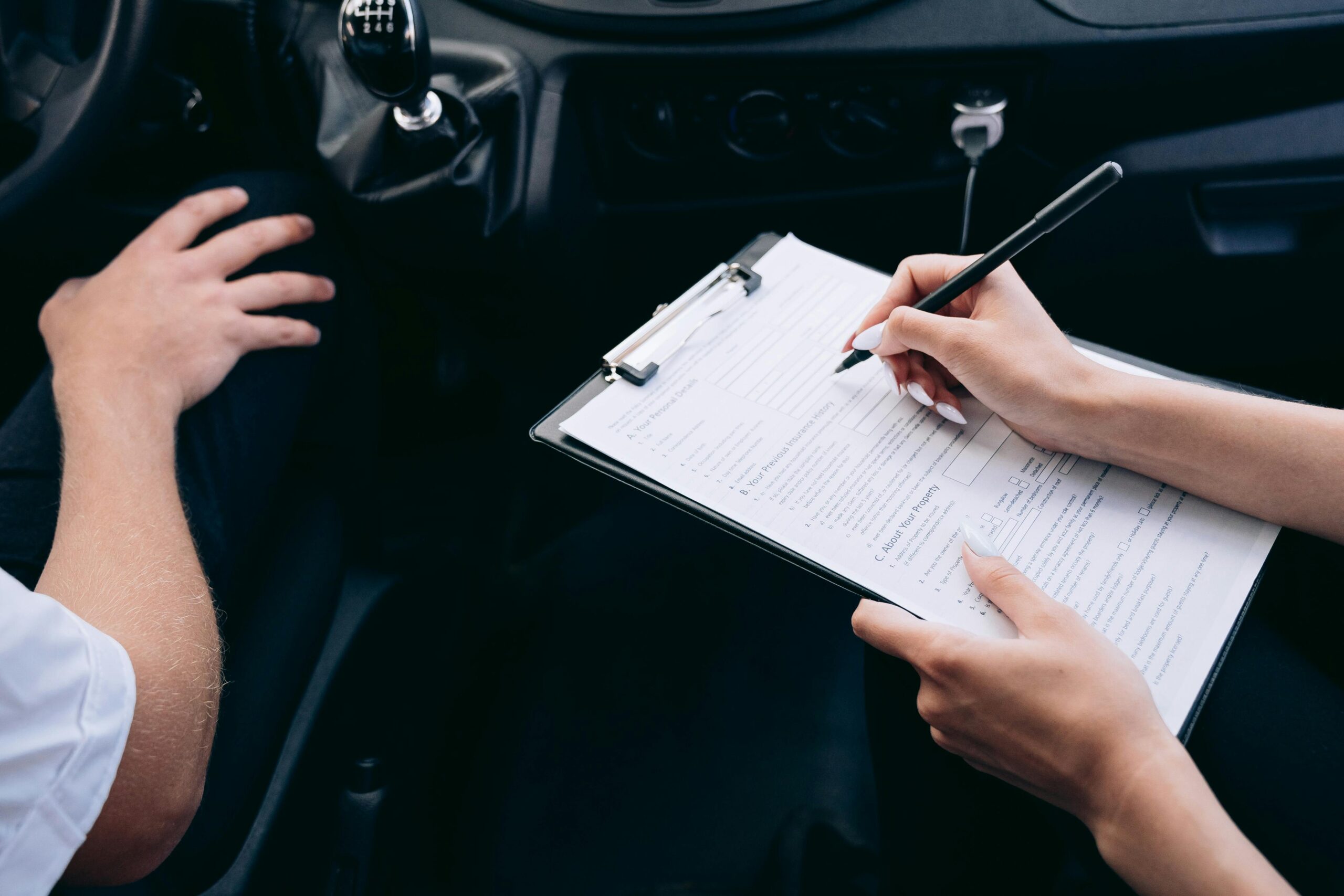 Close-up of a person writing on a clipboard inside a car, showing hands and a gear shift.